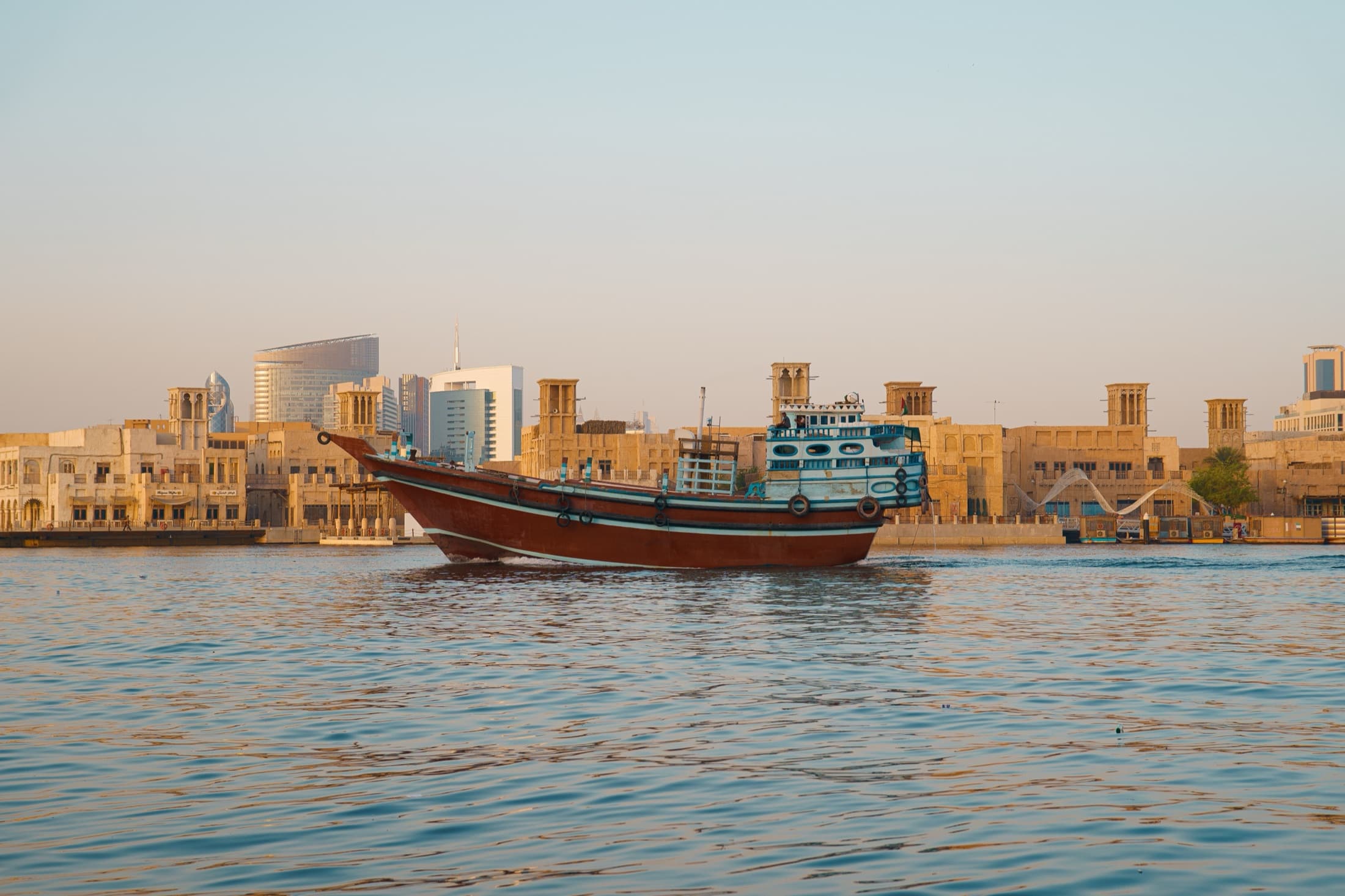 A traditional wooden dhow boat sailing on Dubai Creek with the city's architecture behind it